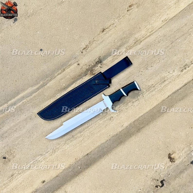 May include: A large, silver-bladed knife with a black and white handle, resting beside its black leather sheath. The knife has a straight edge and a pointed tip. The sheath is rectangular with a strap. The knife and sheath are on a sandy surface.