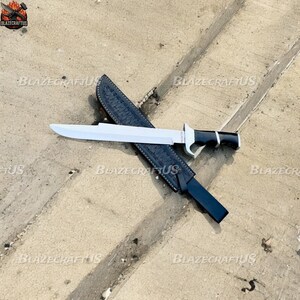 May include: A large, silver blade knife with a black and silver handle, resting on a black leather sheath. The knife has a wide blade and a sturdy handle. The sheath is textured and has a belt loop. The knife is designed for outdoor use.