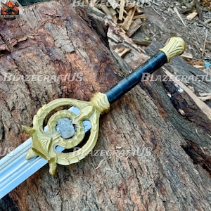 Puede incluir: Una espada decorativa con empuñadura dorada y mango negro. La hoja es plateada y la empuñadura presenta un diseño ornamentado con una piedra central. La espada descansa sobre una superficie de madera texturizada.
