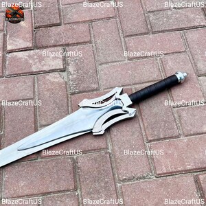 May include: A silver and black sword with a wide, polished blade. The hilt is black with a silver guard. The sword is lying on a brick surface. The sword has a decorative design on the guard.