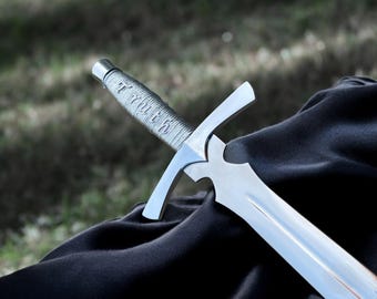 May include: A silver sword with a crossguard and a wrapped handle. The blade is polished and reflects light. The handle has the word "Truth" etched into it. The sword rests on a black surface.