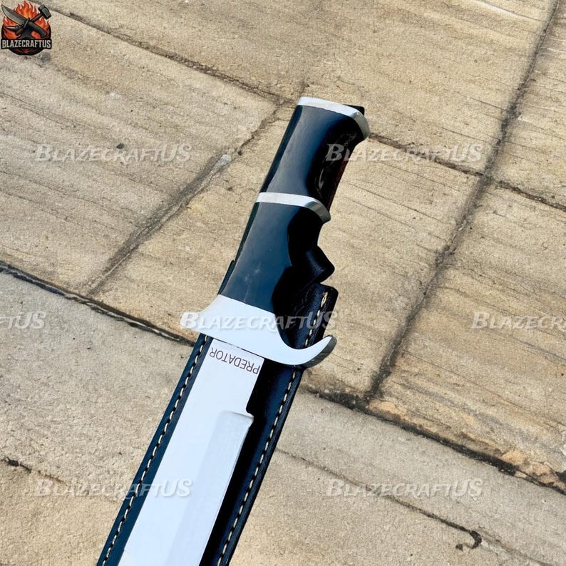 May include: A black and silver knife with the word "PREDATOR" on the sheath. The knife has a black handle and a silver blade. The sheath is black with white accents.