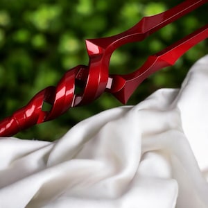 May include: A close-up of a red spear with a twisted central design and sharp, angular tips. The spear is set against a backdrop of white fabric and a blurred green background, highlighting its glossy finish.