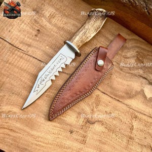 May include: A hunting knife with a serrated blade and a handle made of antler material. The knife is accompanied by a brown leather sheath. The blade has etched text. The knife and sheath are displayed on a wooden surface.