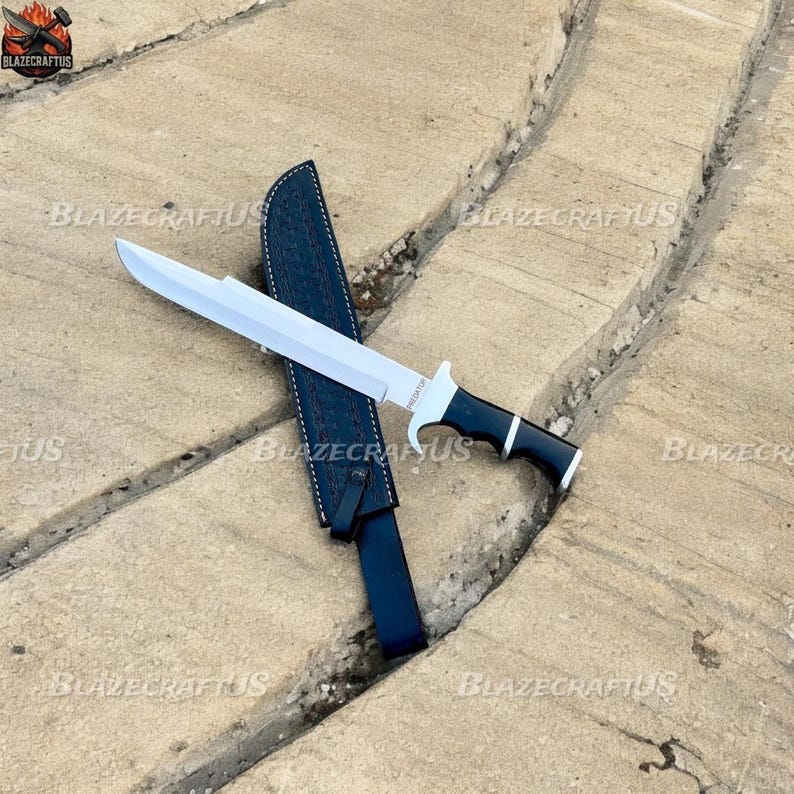 May include: A large, silver blade knife with a black and white handle, resting on a stone surface. The knife is accompanied by a black sheath. The word "PREDATOR" is visible on the blade.
