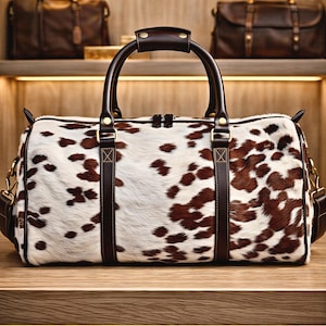 May include: A cowhide duffel bag with a white and brown spotted pattern. The bag has dark brown leather handles, straps, and trim. Two similar leather bags are visible on a shelf in the background.