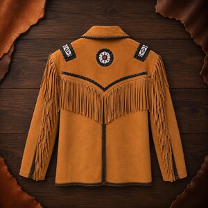 May include: A tan suede jacket featuring long fringe detailing along the sleeves and across the back. The garment is adorned with dark brown braided trim, a circular beaded emblem at the center back, and rectangular beaded patches on the shoulders.