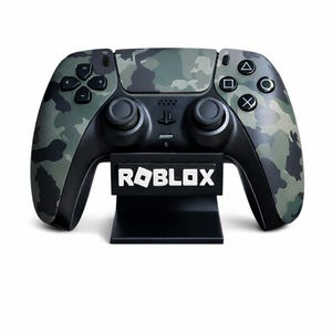 Könnte beinhalten: Ein schwarz-grüner Videospiel-Controller mit Tarnmuster und dem Wort "ROBLOX" in Weiß auf einem schwarzen Ständer. Der Controller hat schwarze Knöpfe und Joysticks. Der Hintergrund ist weiß.