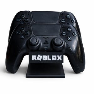 Könnte beinhalten: Zwei schwarze Gamecontroller sind übereinander gestapelt und ruhen auf einem schwarzen Ständer mit dem weißen "ROBLOX"-Logo. Die Controller haben verschiedene Tasten und Joysticks. Der Hintergrund ist weiß.