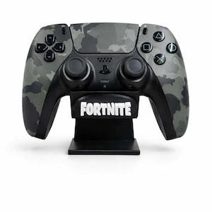 Könnte beinhalten: Ein schwarz-grauer Videospiel-Controller mit Tarnmuster, schwarzen Knöpfen und einem schwarzen Ständer. Auf dem Ständer steht das Wort "FORTNITE" in weißen Blockbuchstaben. Der Controller ist für Spiele konzipiert.