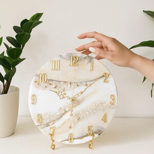 May include: A round wall clock featuring a white and beige marbled resin surface with embedded clear crystal-like fragments. The clock face displays raised gold-coloured numerals from one to twelve and ornate gold-coloured hour and minute hands.