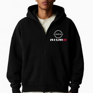 Puede incluir: Sudadera negra con cremallera con el logo "nismo" blanco y rojo y el logo "Nissan" blanco. La sudadera con capucha tiene una capucha con cordón y una cremallera de largo completo. El modelo lleva una camiseta blanca debajo.