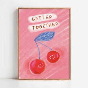 Op de afbeelding: Een ingelijste kunstprint met een roze achtergrond, met twee lachende kersen en een blauw blad. De woorden "BETTER TOGETHER" staan in het wit boven de kersen. Het frame is van hout.
