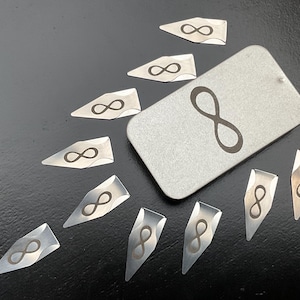 May include: A collection of silver-colored metal charms and a tag, all featuring the infinity symbol. The charms are various shapes, including arrowheads and rectangles. The infinity symbol is etched in a darker tone.