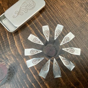 May include: A set of silver guitar picks with a decorative swirl design, arranged in a circle on a dark wooden surface. A small, matching silver tin with a similar design is open nearby. The picks are approximately 1 inch long.