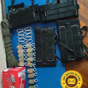 May include: A collection of black straps, metal carabiners, and hardware components arranged on a blue mat. Includes a coil of green rope and a red box with the text "ALKO". A yellow sticker with the text "ASK BEFORE BUYING" is also visible.