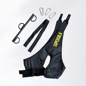 May include: Black exercise harness with the text "SUPERSILA" in yellow, a black bar with rings, two black straps, and three silver carabiner clips. The harness features adjustable straps and a buckle.