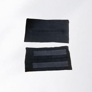 May include: Two black fabric wraps, likely for a wrist or ankle, are displayed against a white background. One wrap is plain, while the other has two hook-and-loop fastener strips for secure closure. The wraps appear to be made of a soft, flexible material.