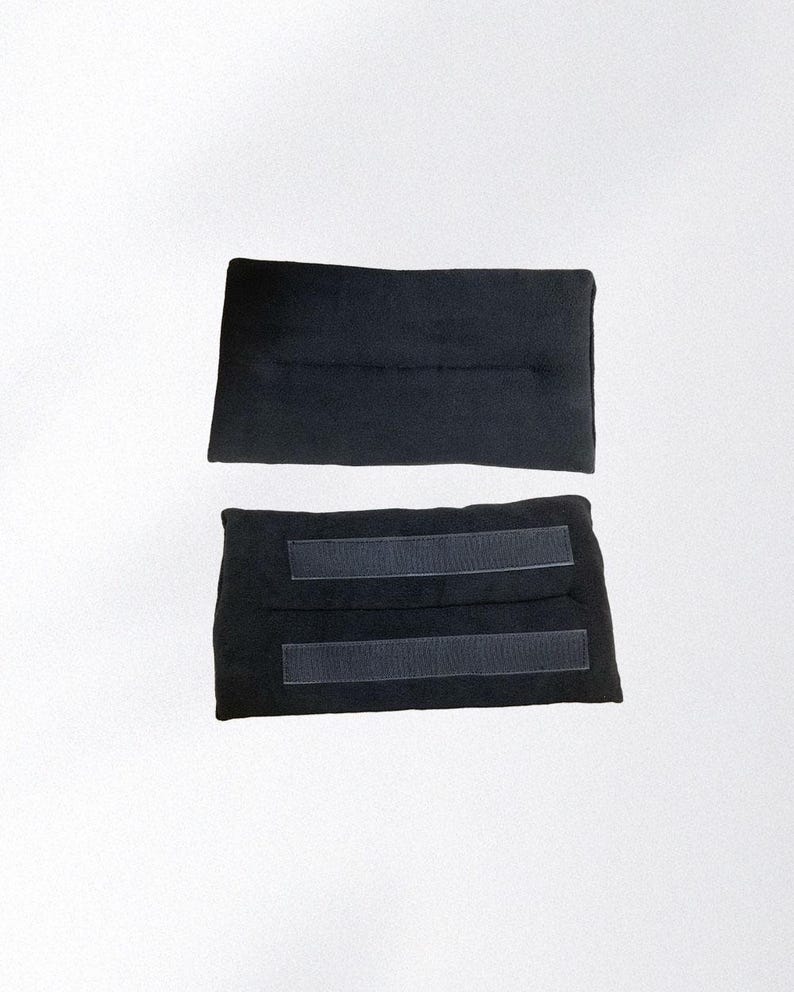 May include: Two black fabric wraps, likely for a wrist or ankle, are displayed against a white background. One wrap is plain, while the other has two hook-and-loop fastener strips for secure closure. The wraps appear to be made of a soft, flexible material.
