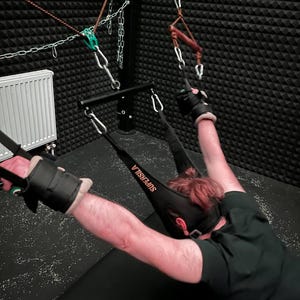 May include: A black and gray bondage sling with the word "SUPERSILA" printed on it. The sling is attached to a metal frame with chains and pulleys. Black wrist cuffs are also visible.