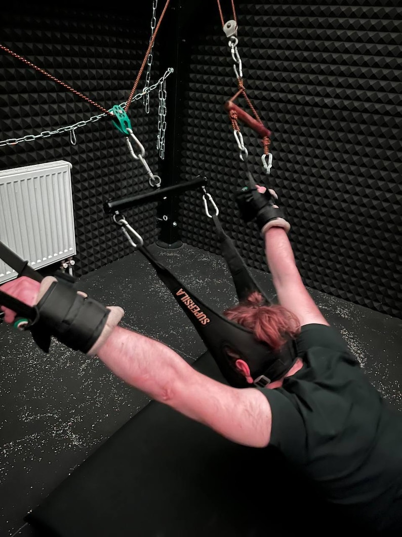 May include: A black and gray bondage sling with the word "SUPERSILA" printed on it. The sling is attached to a metal frame with chains and pulleys. Black wrist cuffs are also visible.