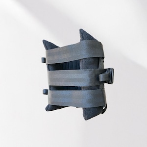 May include: A black, padded item with three gray straps and a black buckle. The item has a unique shape with two pointed sections at the top and bottom. The straps are secured with black plastic buckles.