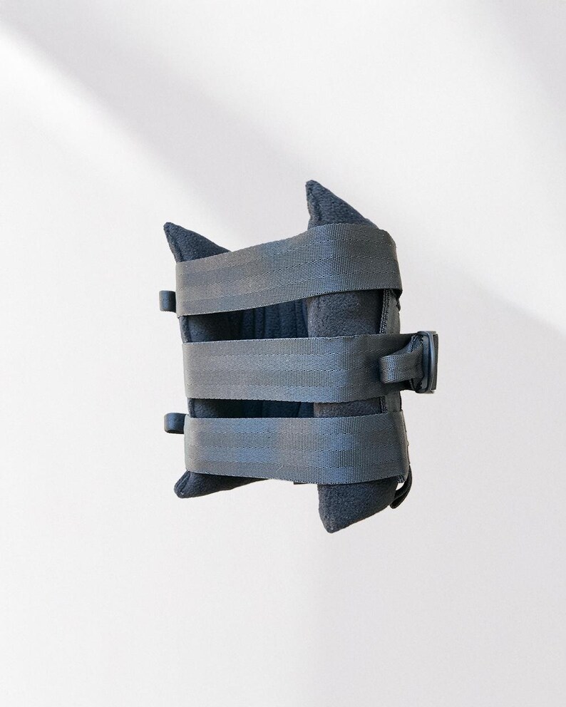 May include: A black, padded item with three gray straps and a black buckle. The item has a unique shape with two pointed sections at the top and bottom. The straps are secured with black plastic buckles.