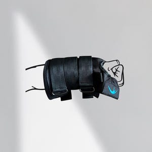 May include: Black wrist brace with adjustable straps and a blue logo. The brace is designed to support the wrist and hand. The image shows the brace in a fist position, suggesting its use in sports or injury recovery.