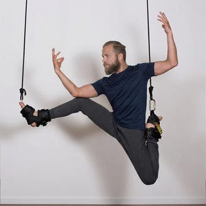 May include: A person suspended in the air, wearing black ankle straps attached to ropes. The person is wearing a navy blue t-shirt and grey sweatpants, demonstrating a yoga pose. The straps have yellow text that reads "SUPERSILA".