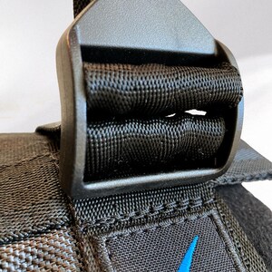May include: Close-up of a black nylon strap with a plastic buckle. The strap is woven with a textured pattern. A blue swoosh logo is visible on the strap. The buckle is a matte black color.
