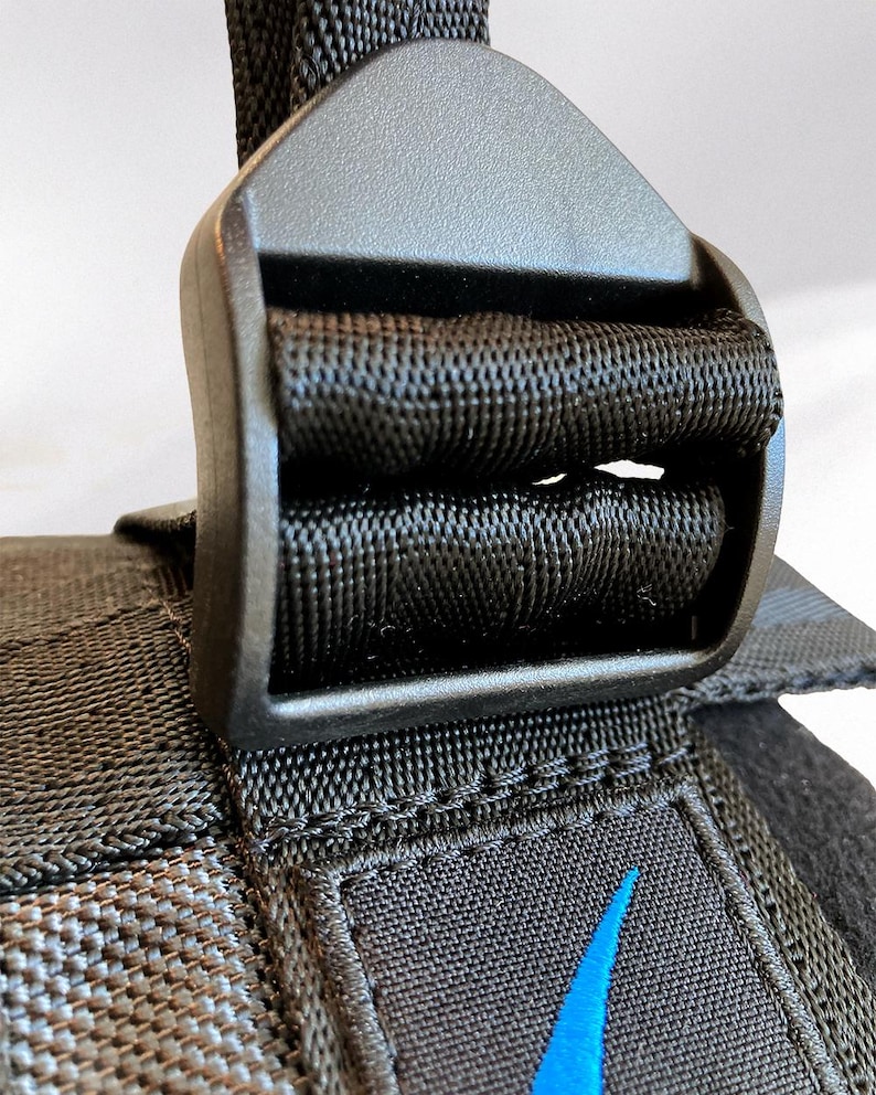 May include: Close-up of a black nylon strap with a plastic buckle. The strap is woven with a textured pattern. A blue swoosh logo is visible on the strap. The buckle is a matte black color.