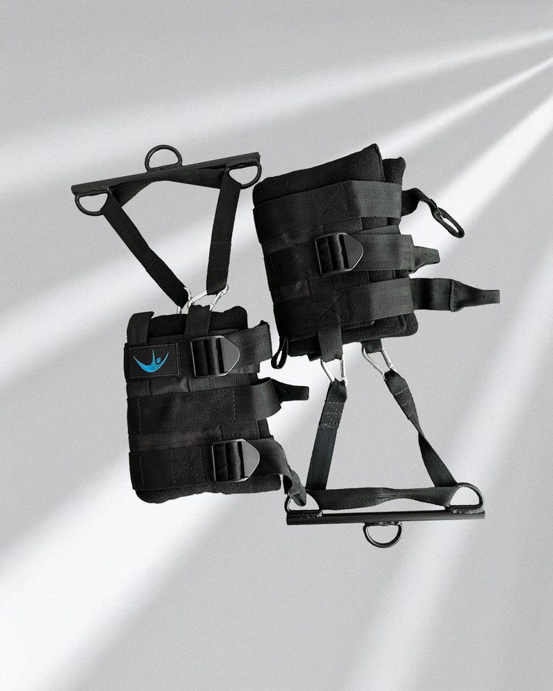 May include: Black inversion boots with multiple adjustable straps and metal D-rings. The boots have a padded interior and a blue logo patch. The design includes a metal bar with a D-ring for hanging. The boots are designed for inversion therapy.