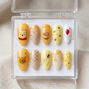 May include: A set of press-on nails in a clear plastic case. The nails feature yellow, beige, and white designs with Winnie the Pooh, honey, bees, and a red balloon. The nails are oval-shaped.