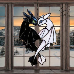 Puede incluir: Vidriera con dos dragones, uno negro con ojos verdes y otro blanco con ojos azules, enfrentados en forma de corazón. La obra cuelga de una cadena frente a una ventana con un paisaje invernal de fondo.