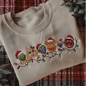 Op de afbeelding: Een beige sweater met ronde hals en een borduursel van zes Muppet-figuren met kerstmutsen. De figuren staan op een rij en zijn verbonden door een kleurrijke kerstverlichting die over de voorkant van het kledingstuk gedrapeerd is.