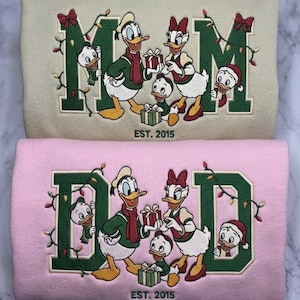 Puede incluir: Dos sudaderas dobladas, una beige y otra rosa, con bordados de Donald, Daisy y patitos con luces navideñas. La sudadera beige muestra el texto M-M y la rosa D-D, ambas con la inscripción EST. 2015 en la parte inferior.