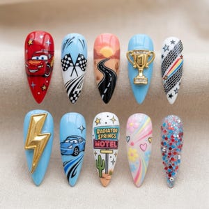 Puede incluir: Un conjunto de diez uñas artificiales largas en forma de almendra con varios diseños. Los diseños incluyen un coche rojo, banderas a cuadros, una carretera desértica, un trofeo dorado, huellas de neumáticos, un rayo dorado, un coche azul y el texto "RADIATOR SPRINGS MOTEL".