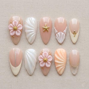 Puede incluir: Un conjunto de diez uñas artificiales en forma de almendra con varios diseños. Las uñas presentan una base rosa claro con adornos como una estrella de mar dorada, una flor rosa y diseños de conchas blancas.