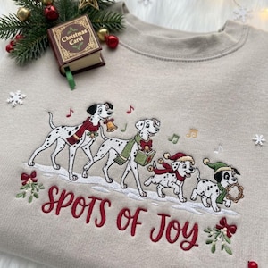 May include: A beige crewneck sweatshirt featuring an embroidered design of four Dalmatian dogs wearing scarves and hats, singing with musical notes. The text "SPOTS of Joy" is embroidered in red below the dogs, flanked by sprigs of mistletoe.