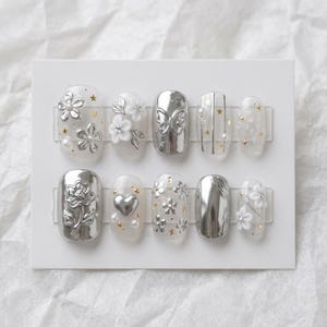 Könnte beinhalten: Ein Set aus zehn künstlichen Nägeln mit verschiedenen Designs. Die Nägel weisen silbermetallische und transparente Designs mit floralen, Schmetterlings- und Sternakzenten auf. Einige Nägel haben Perlen- und Goldverzierungen. Die Nägel werden auf einer weißen Karte präsentiert.