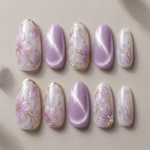 Pode incluir: Um conjunto de dez unhas artificiais em vários formatos ovais. As unhas apresentam um design de flores lavanda com contornos e detalhes dourados. Algumas unhas têm um acabamento perolado, enquanto outras são de cor lavanda sólida.