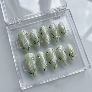 May include: A set of ten oval-shaped, press-on nails in a clear plastic case. The nails are a pale green with white floral designs, dark green stems, and gold or clear gem accents. The nails are designed for cosmetic use.