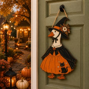 May include: A wooden door hanger shaped like a goose wearing a black witch hat with orange trim and a floral accent. The goose wears a black cape over an orange dress featuring a small black cat graphic and a pumpkin-shaped button at the neck.