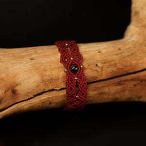 Op de afbeelding: Een rood en gouden gevlochten armband met een zwarte edelsteen in het midden.