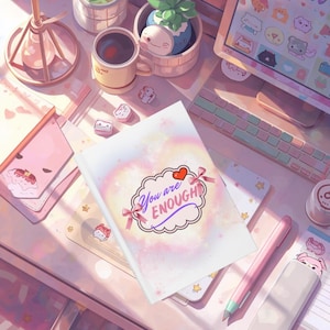 May include: A notebook with the text "You are ENOUGH" in a decorative cloud-shaped frame, accented with pink bows and a red heart. The notebook is on a desk with other stationery and a computer.