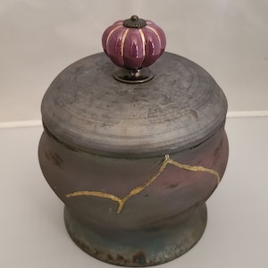 May include: A ceramic lidded jar with a purple and gold design. The lid has a purple knob in the shape of a pumpkin.