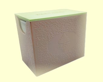 Pode incluir: Uma caixa de lenços quadrada com tampa verde claro e corpo rosa pálido. O corpo tem um padrão em relevo texturizado de folhas e uma forma de espadas no centro. A caixa de lenços está em um fundo amarelo claro.