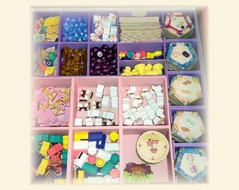 Pode incluir: Um organizador de jogos de tabuleiro rosa e roxo cheio de peças de jogo. Os compartimentos contêm fichas de madeira coloridas, cartas e ladrilhos hexagonais. As peças são em tons de azul, amarelo, castanho e rosa.
