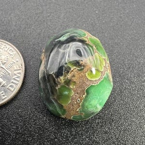 Op de afbeelding: Een groene, zwarte en bruine cabochon edelsteen met een glad, gepolijst oppervlak. De steen heeft een uniek patroon van swirls en vlekken.