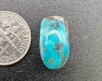Puede incluir: Un cabujón turquesa con un color azul verdoso y matriz marrón y negra. El cabujón tiene forma ovalada y una superficie lisa y pulida.